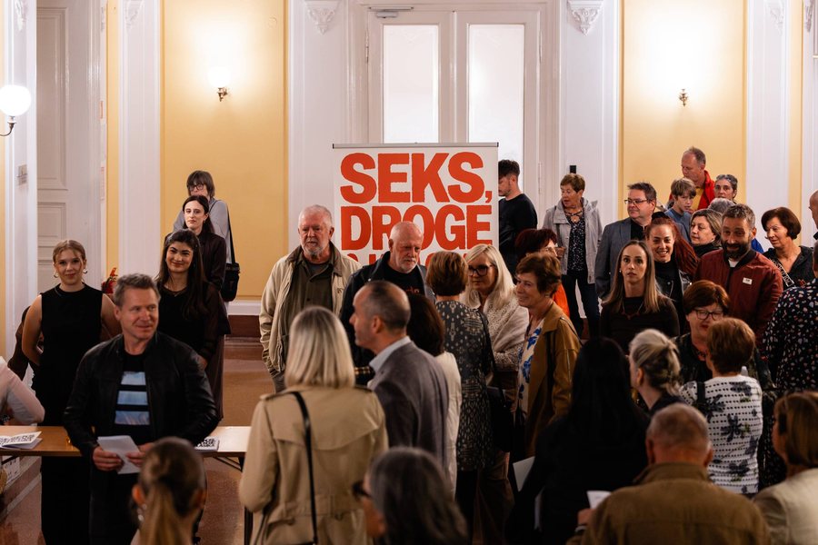 Skupina obiskovalcev stoji pred promocijskim napisom predstave Seks, droge & klasika.